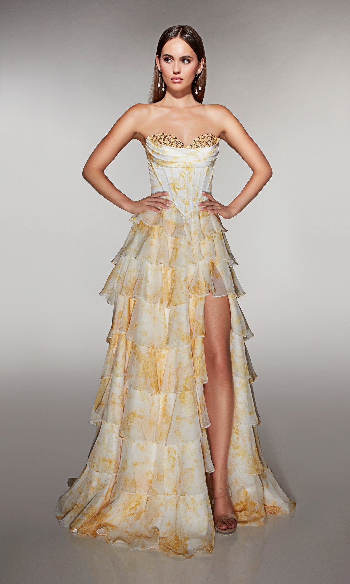 A woman wears the Alyce 62192 long prom dress in yellow/white, featuring a strapless corset bodice, gold details, a tiered chiffon ruffle skirt with thigh-high slit, and poses against a neutral background.
